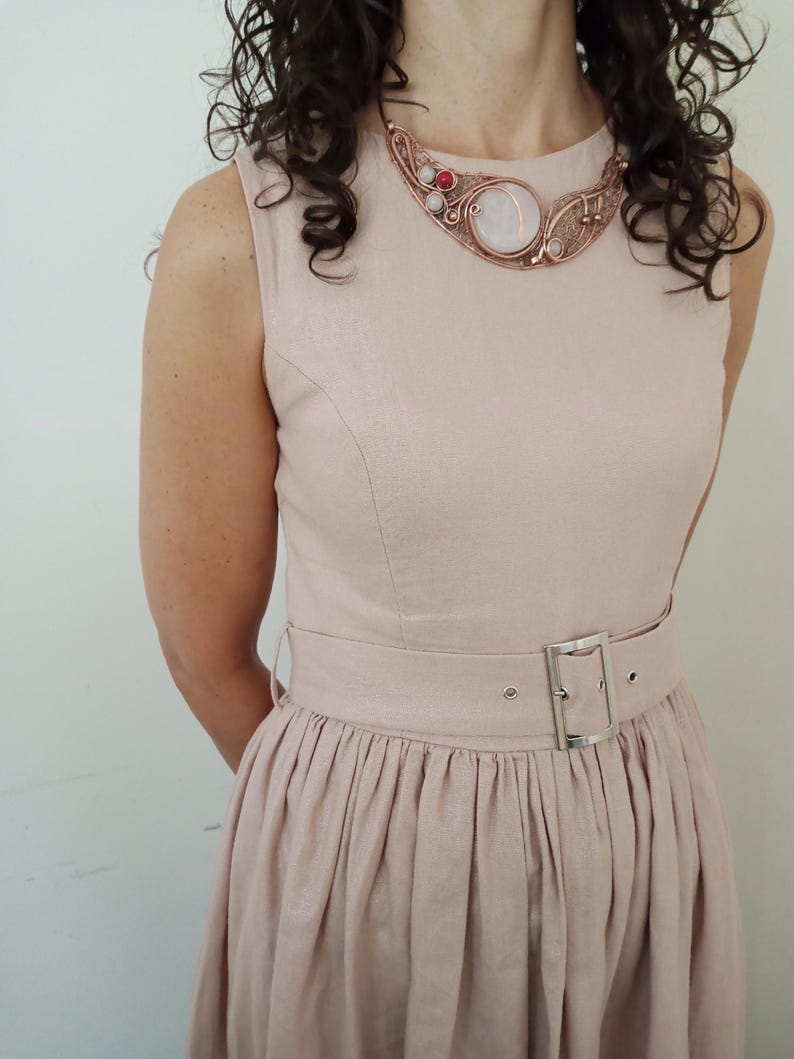 May include: A sleeveless light beige dress with a matching belt featuring a silver buckle. The dress is paired with a copper wire-wrapped statement necklace that includes a large central white stone, smaller white beads, and a single red bead.