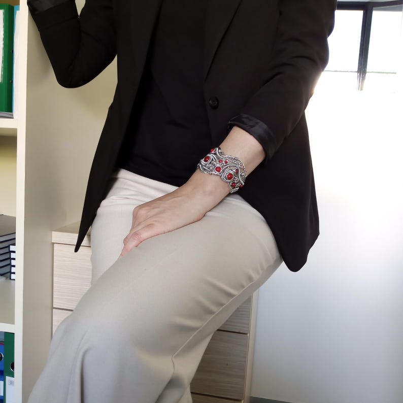 May include: A silver cuff bracelet with red gemstone accents. The bracelet features an ornate, filigree design. The person in the image is wearing a black blazer, black shirt, and tan pants.