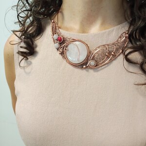 May include: A copper wire-wrapped necklace featuring a large, round, translucent white stone centerpiece. The intricate metalwork includes smaller white and red spherical beads integrated into the design, creating a wide, curved collar shape.