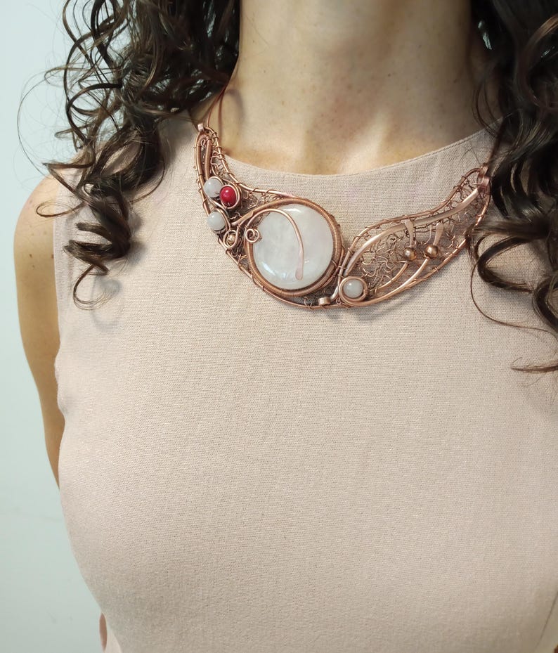 May include: A copper wire-wrapped necklace featuring a large, round, translucent white stone centerpiece. The intricate metalwork includes smaller white and red spherical beads integrated into the design, creating a wide, curved collar shape.