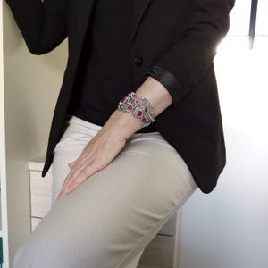 May include: A silver-toned cuff bracelet with red gemstone accents. The bracelet features an intricate, openwork design with multiple red stones. The person is wearing a black blazer, black top, and khaki pants.