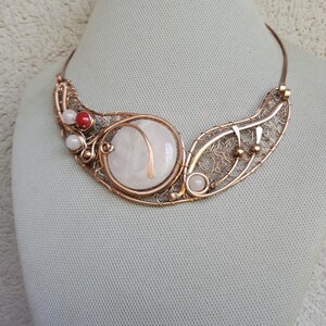 May include: A copper wire-wrapped necklace featuring a large, round, white stone centerpiece, accented with smaller white beads and a single red bead. The design incorporates intricate wire filigree patterns and a curved copper wire frame.