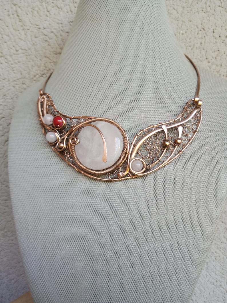 May include: A copper wire-wrapped necklace featuring a large, round, white stone centerpiece, accented with smaller white beads and a single red bead. The design incorporates intricate wire filigree patterns and a curved copper wire frame.