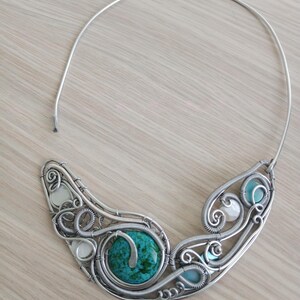 May include: A handcrafted silver wire necklace with a large turquoise stone and smaller white and blue stones. The necklace has a unique, artistic design with swirling wire patterns and a simple, curved neckband.
