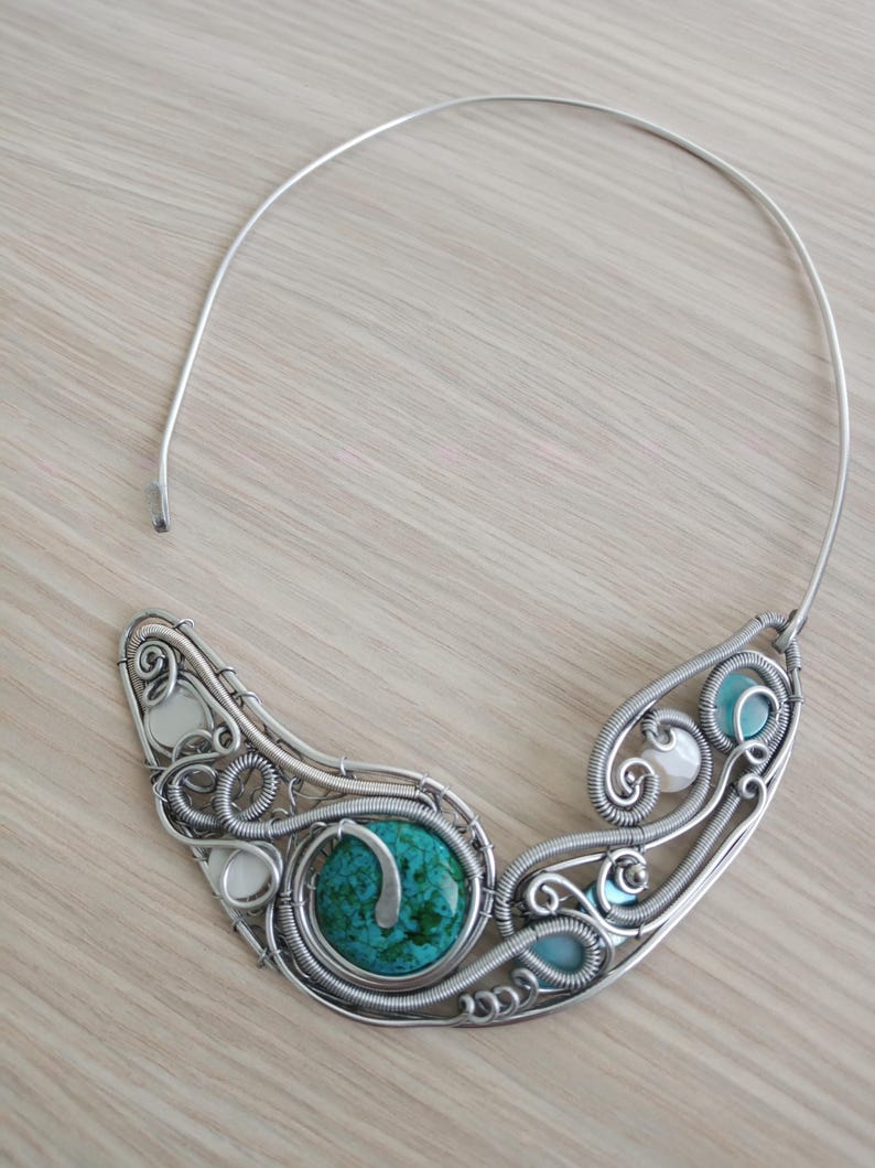 May include: A handcrafted silver wire necklace with a large turquoise stone and smaller white and blue stones. The necklace has a unique, artistic design with swirling wire patterns and a simple, curved neckband.
