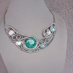 May include: A silver-coloured necklace with a central turquoise stone and an elaborate wire-wrapped design. The necklace incorporates smaller turquoise and white stones, creating a unique, handcrafted piece.
