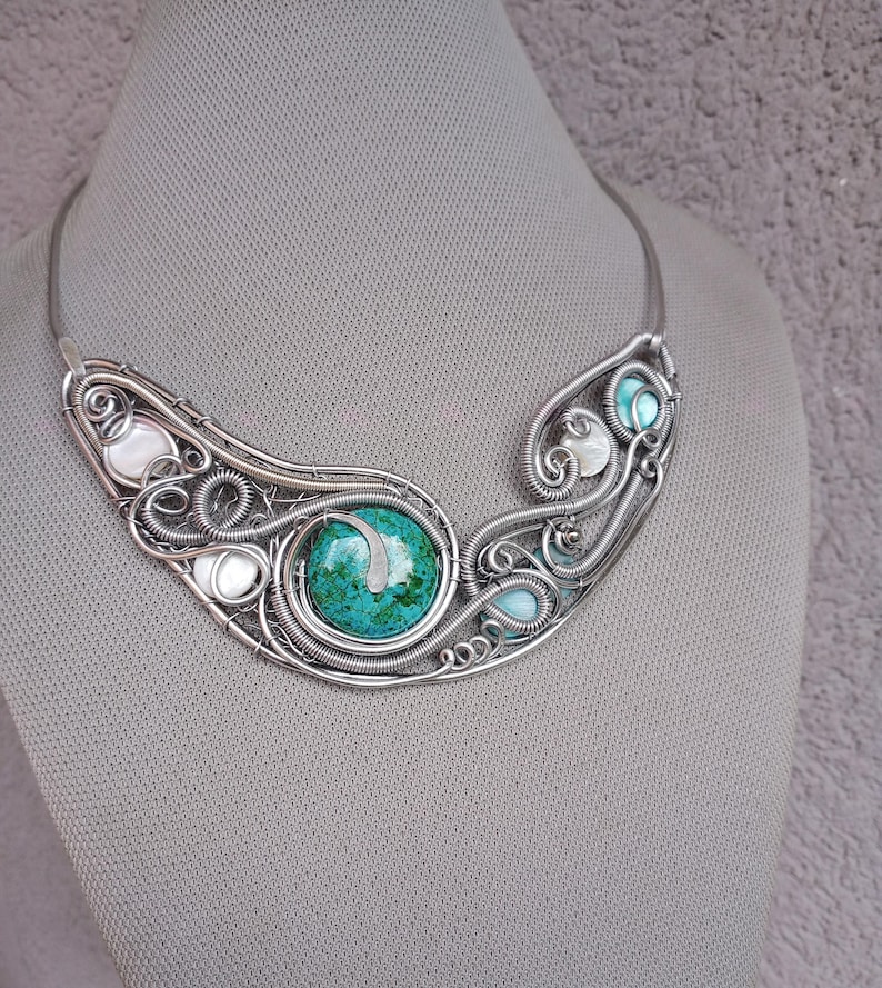 May include: A silver-coloured necklace with a central turquoise stone and an elaborate wire-wrapped design. The necklace incorporates smaller turquoise and white stones, creating a unique, handcrafted piece.