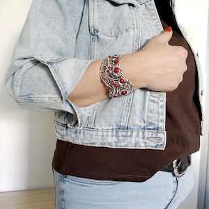 May include: A silver-toned cuff bracelet with intricate wirework and vibrant red gemstone accents. The bracelet is a statement piece, featuring a detailed design with multiple red stones. The jewelry is worn on a wrist, showcasing its craftsmanship and bold aesthetic.