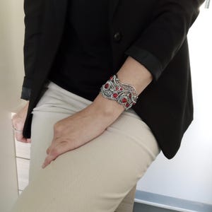 May include: A silver-toned cuff bracelet with red gemstone accents. The bracelet features an openwork design with intricate patterns and is adorned with multiple round red stones. The jewelry is worn on a person's wrist.