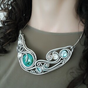 May include: A handcrafted silver necklace with a large turquoise stone and smaller blue and white stones. The wire-wrapped design features intricate swirls and curves. The necklace is a statement piece, with a unique, artistic design.