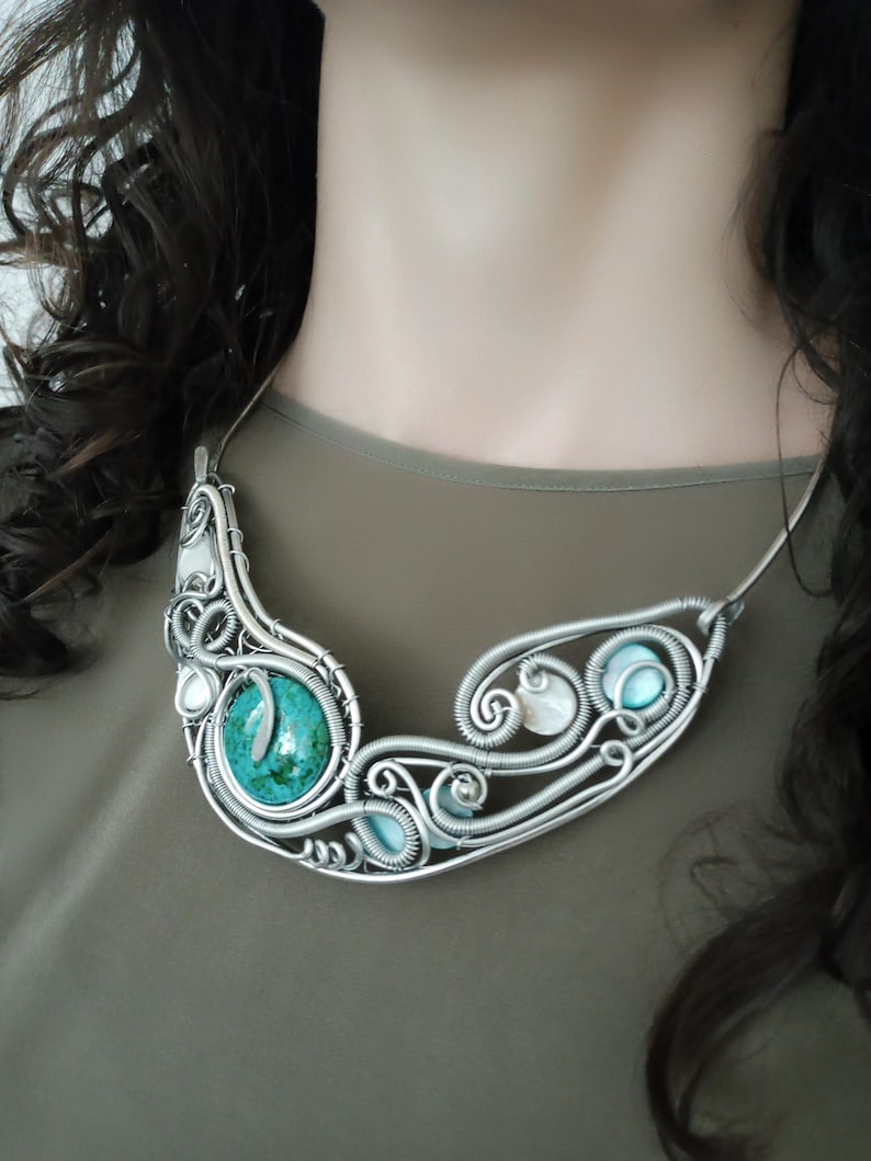 May include: A handcrafted silver necklace with a large turquoise stone and smaller blue and white stones. The wire-wrapped design features intricate swirls and curves. The necklace is a statement piece, with a unique, artistic design.