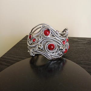 May include: A silver-toned wire-wrapped cuff bracelet with embedded red gemstones. The bracelet features intricate wirework with swirling patterns and is adorned with several round, red stones. The jewelry piece is displayed on a dark surface.