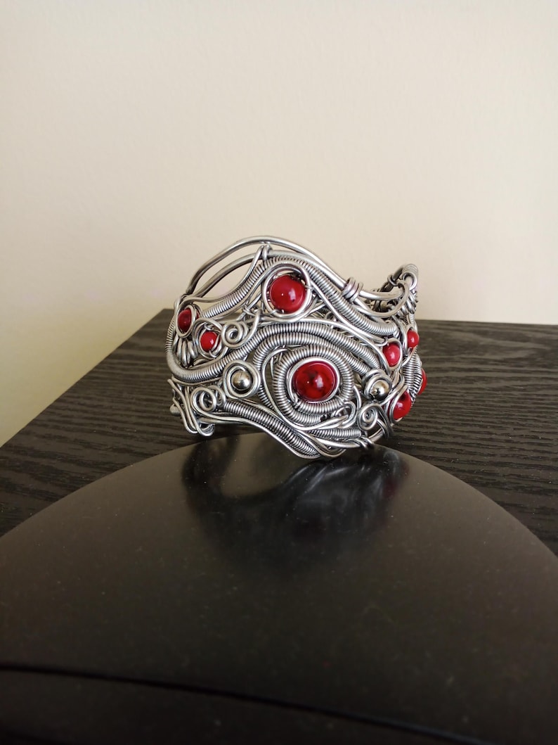 May include: A silver-toned wire-wrapped cuff bracelet with embedded red gemstones. The bracelet features intricate wirework with swirling patterns and is adorned with several round, red stones. The jewelry piece is displayed on a dark surface.