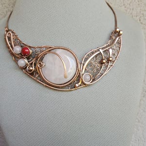 May include: A copper wire-wrapped necklace featuring a large, round, pale pink stone centerpiece. The design includes intricate wire mesh detailing, small white beads, and a single vibrant red bead, all set within a curved, metallic copper frame.