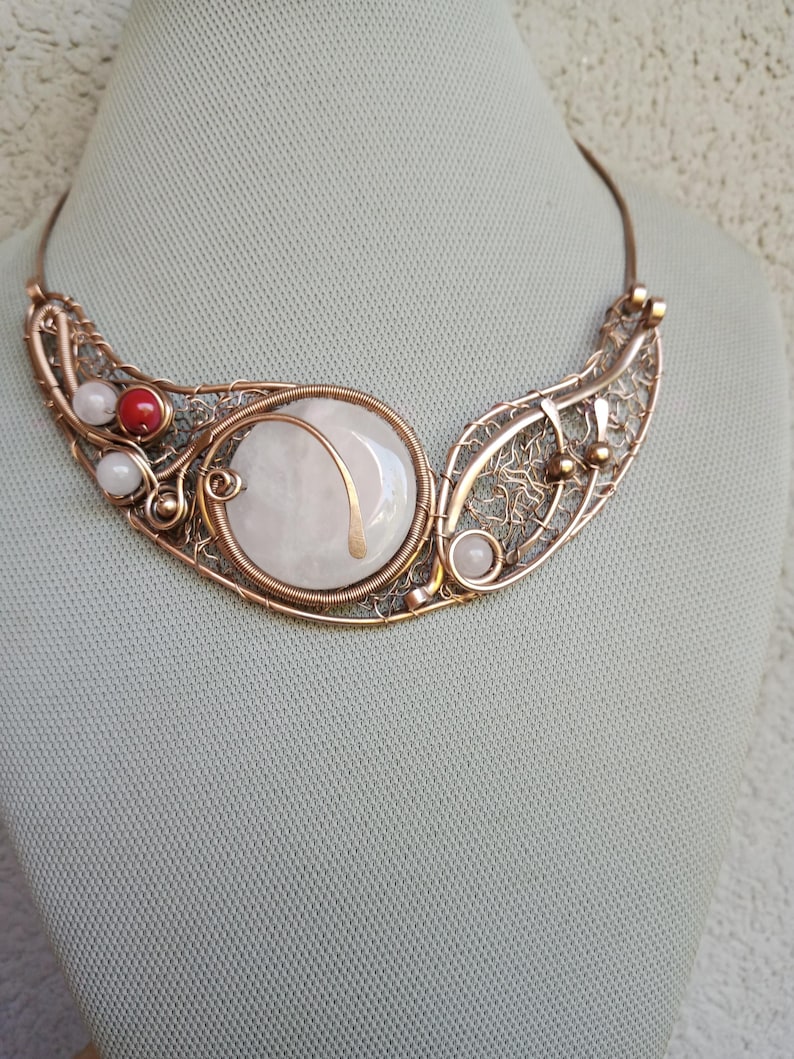 May include: A copper wire-wrapped necklace featuring a large, round, pale pink stone centerpiece. The design includes intricate wire mesh detailing, small white beads, and a single vibrant red bead, all set within a curved, metallic copper frame.