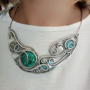 May include: A handcrafted silver necklace with a large, round turquoise stone and smaller turquoise and white stones. The wire-wrapped design features intricate swirls and curves. The necklace is worn around the neck, showcasing its unique, artistic style.