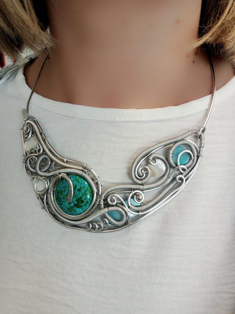 May include: A handcrafted silver necklace with a large, round turquoise stone and smaller turquoise and white stones. The wire-wrapped design features intricate swirls and curves. The necklace is worn around the neck, showcasing its unique, artistic style.