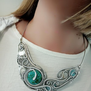 May include: A silver wire-wrapped necklace with turquoise and light blue stones. The necklace has an intricate, swirling design with a central turquoise stone and smaller blue stones. The necklace is worn against a white top.