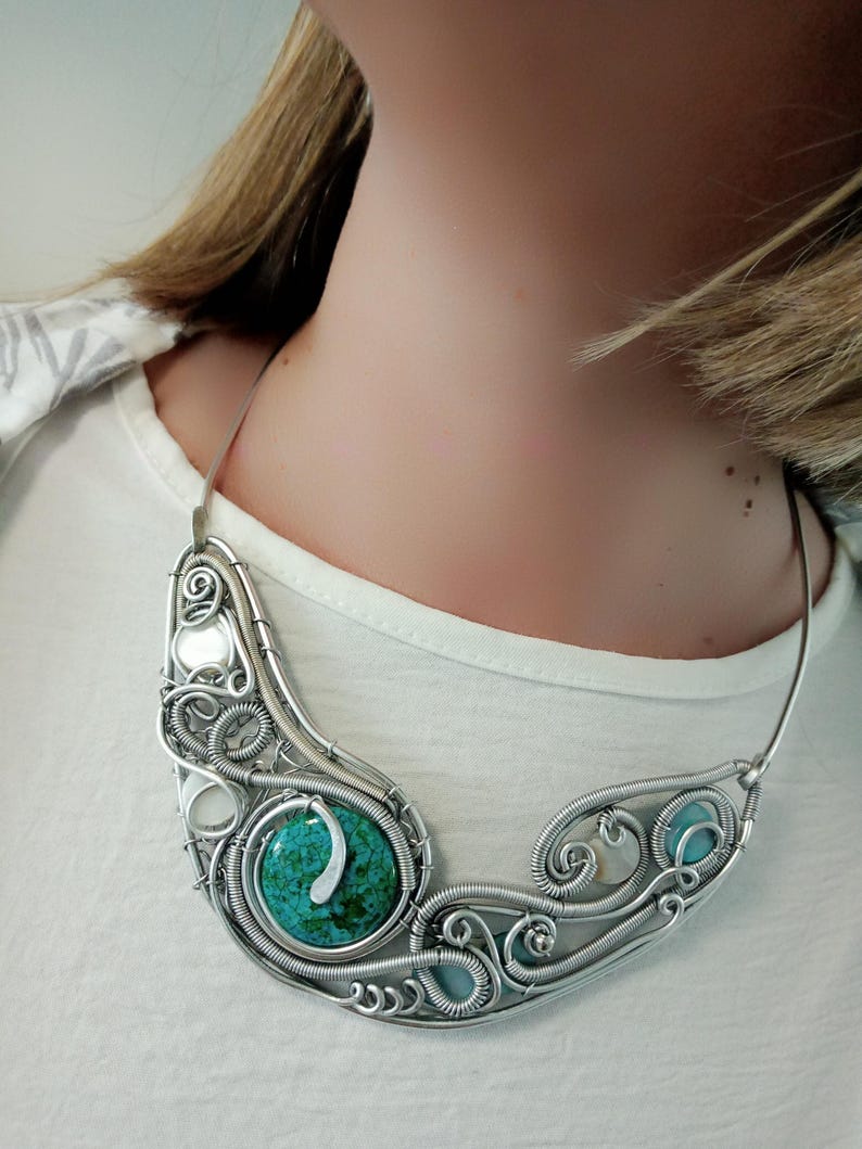 May include: A silver wire-wrapped necklace with turquoise and light blue stones. The necklace has an intricate, swirling design with a central turquoise stone and smaller blue stones. The necklace is worn against a white top.