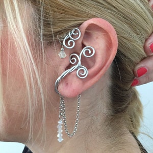 May include: Silver ear cuff with a delicate chain and crystal beads dangling from it. The cuff is shaped like a branch with multiple curls.