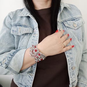 May include: A silver-toned cuff bracelet with red stone accents. The bracelet features an intricate, openwork design with circular and leaf-like elements. The red stones are evenly spaced throughout the bracelet.