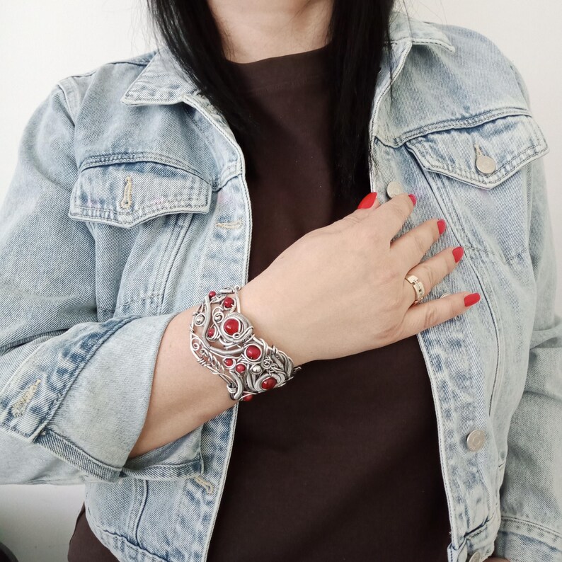 May include: A silver-toned cuff bracelet with red stone accents. The bracelet features an intricate, openwork design with circular and leaf-like elements. The red stones are evenly spaced throughout the bracelet.