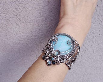 May include: A silver wire wrapped bracelet with a large, light blue gemstone in the center. The gemstone is surrounded by smaller, iridescent gemstones.