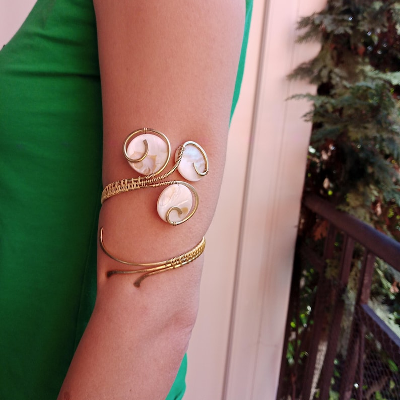 Op de afbeelding: Goudkleurige armmanchet met drie cirkelvormige, iriserende schelpaccenten. De manchet wikkelt zich om de bovenarm met een delicaat, met draad omwikkeld ontwerp. Het sieraad wordt gedragen op de arm van een persoon, tegen een groene top.