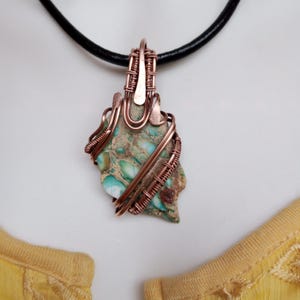 May include: A handcrafted pendant featuring a natural stone with shades of green, brown, and white, wrapped in copper wire. The pendant hangs from a black leather cord, showcasing a unique, earthy design. The stone's irregular shape adds to its organic appeal.