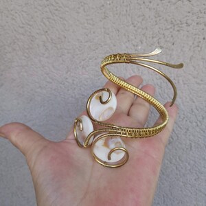 Op de afbeelding: Goudkleurige armmanchet met drie ovale, witte en bruine schelpaccenten. De manchet heeft een geweven ontwerp en gebogen, open uiteinden. De sieraden worden in een hand gehouden en tonen de ingewikkelde details en het vakmanschap van het stuk.