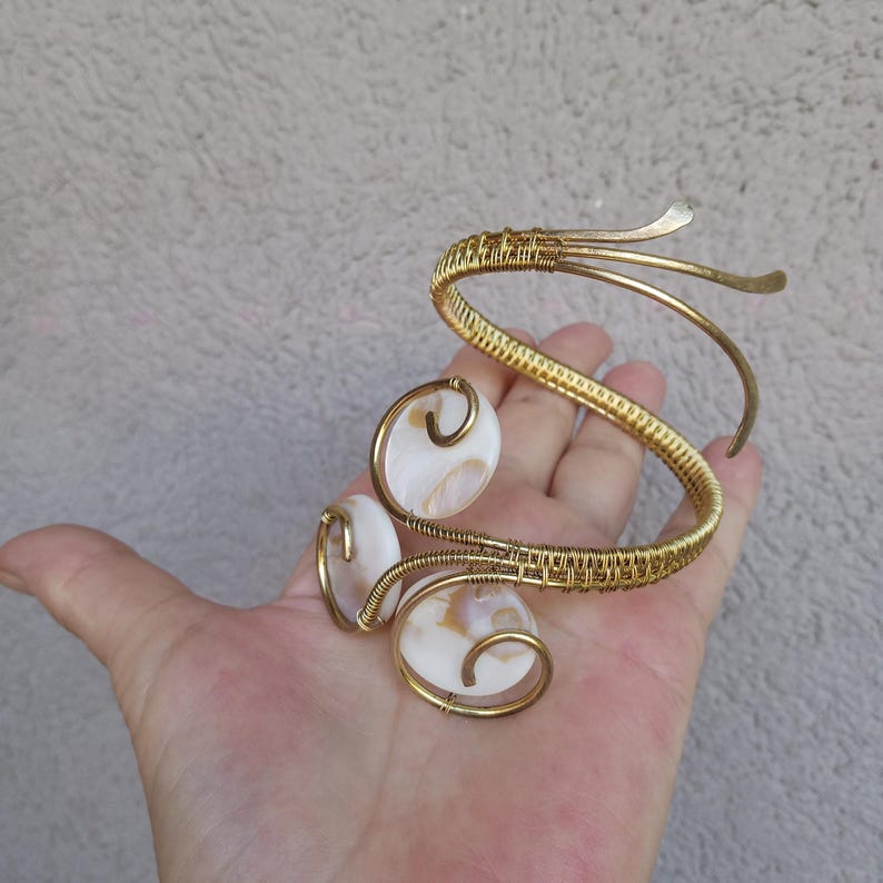 Op de afbeelding: Goudkleurige armmanchet met drie ovale, witte en bruine schelpaccenten. De manchet heeft een geweven ontwerp en gebogen, open uiteinden. De sieraden worden in een hand gehouden en tonen de ingewikkelde details en het vakmanschap van het stuk.
