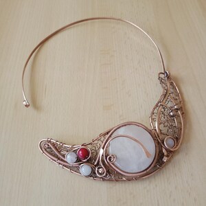 May include: A copper wire torque necklace featuring a large circular white stone, a smaller red bead, and two small white beads. The metalwork consists of intricate coiled and woven copper wire forming a crescent shape along the collar.