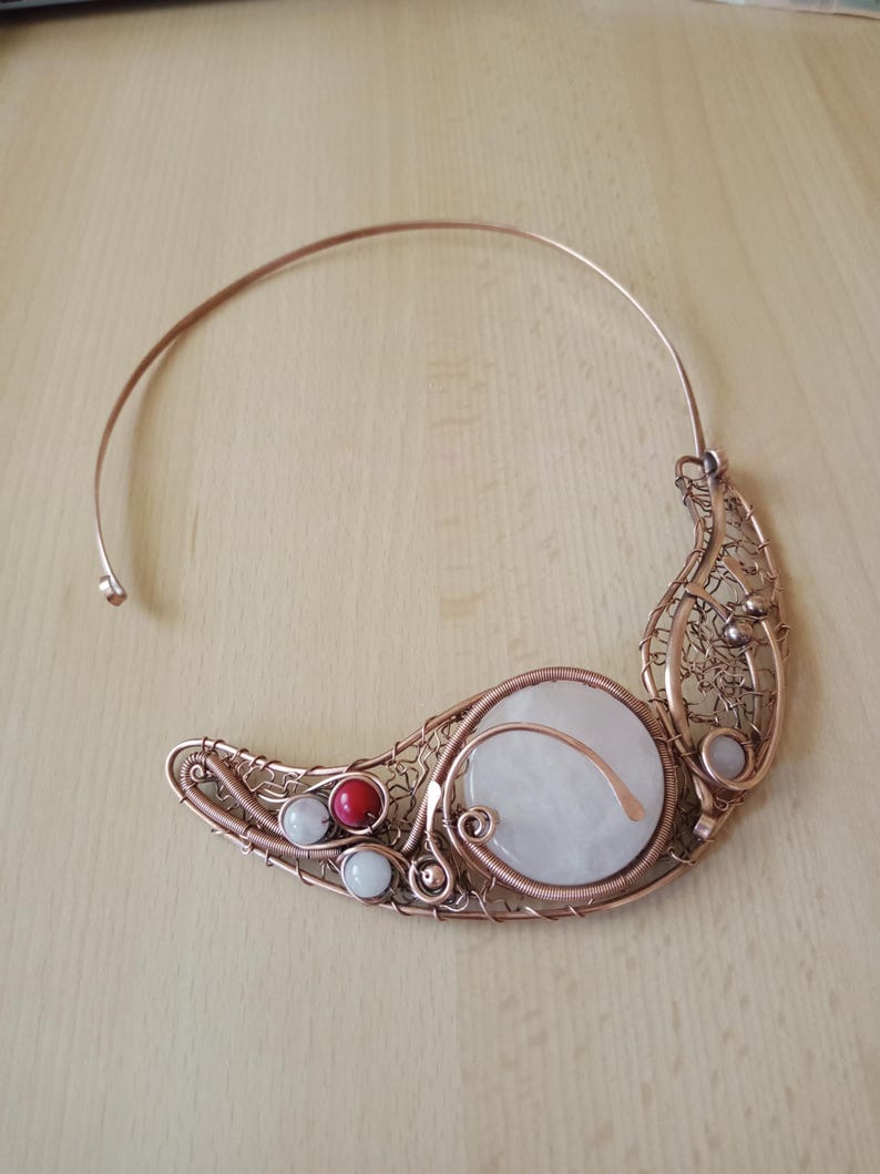 May include: A copper wire torque necklace featuring a large circular white stone, a smaller red bead, and two small white beads. The metalwork consists of intricate coiled and woven copper wire forming a crescent shape along the collar.