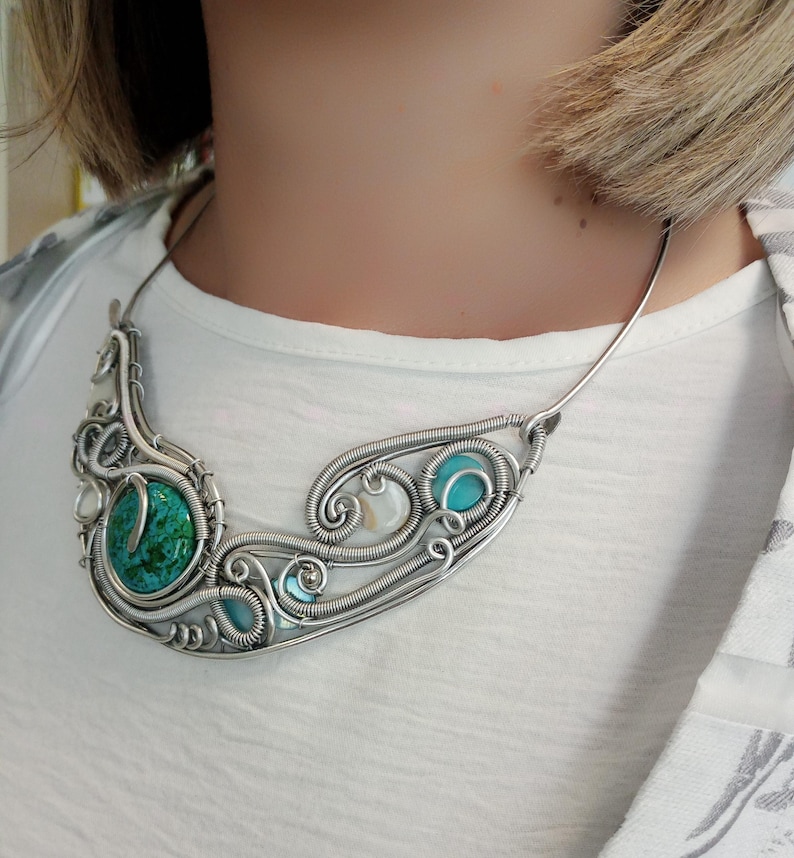 May include: A handcrafted silver wire necklace with a large, round, turquoise-coloured stone and smaller turquoise and white stones. The necklace has a unique, swirling design.
