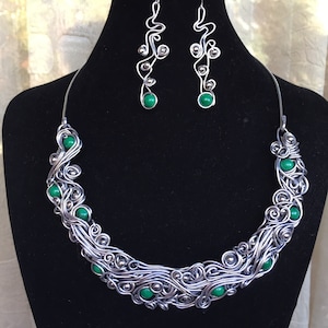 K&ouml;nnte beinhalten: Ein Halskette und Ohrringe aus Silberdraht mit gr&uuml;nen Edelstein-Akzenten. Die Halskette ist ein Choker-Stil mit einem wirbelnden Drahtdesign. Die Ohrringe sind lang und h&auml;ngen mit einem &auml;hnlichen wirbelnden Drahtdesign.