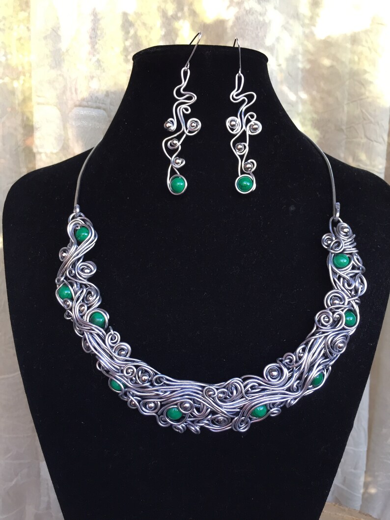 K&ouml;nnte beinhalten: Ein Halskette und Ohrringe aus Silberdraht mit gr&uuml;nen Edelstein-Akzenten. Die Halskette ist ein Choker-Stil mit einem wirbelnden Drahtdesign. Die Ohrringe sind lang und h&auml;ngen mit einem &auml;hnlichen wirbelnden Drahtdesign.