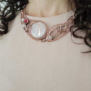 May include: A copper wire-wrapped necklace featuring a large, round, milky white cabochon stone with a copper inlay. The design includes intricate wire filigree, small round white beads, and a single vibrant red bead set within the copper frame.