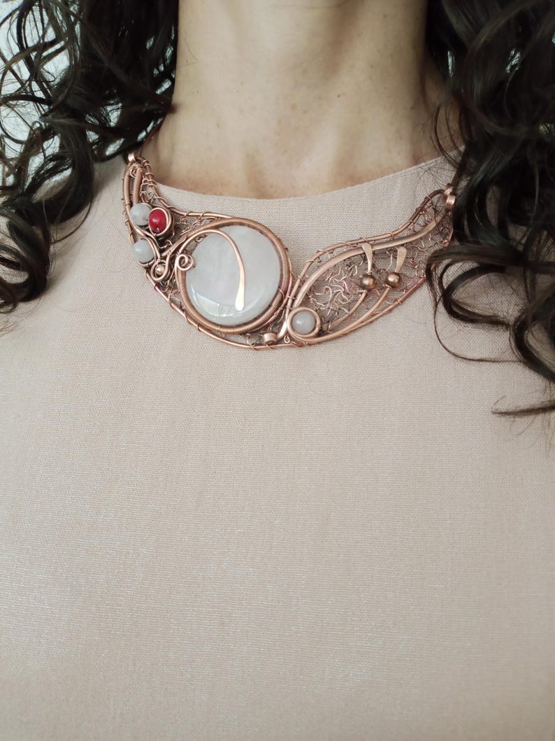May include: A copper wire-wrapped necklace featuring a large, round, milky white cabochon stone with a copper inlay. The design includes intricate wire filigree, small round white beads, and a single vibrant red bead set within the copper frame.
