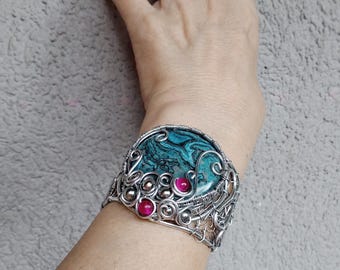 May include: A silver-toned wire-wrapped cuff bracelet. The bracelet features a large, oval-shaped stone with swirling teal and black patterns. Accents include small, round, pink stones. The intricate wirework adds a decorative touch to this unique jewelry piece.
