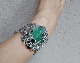 May include: A silver-toned wire-wrapped cuff bracelet featuring a large, oval, green gemstone as the focal point. The bracelet incorporates black and gray accent stones and intricate wirework detailing. The bracelet is worn on a wrist.
