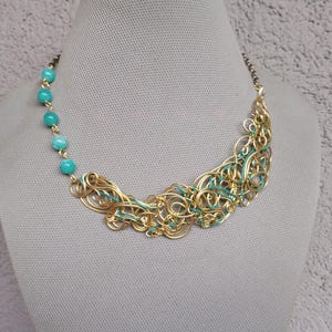 May include: A statement necklace with a gold-coloured wire design and turquoise beads. The intricate wirework is interwoven with teal accents. The necklace is displayed on a mannequin, showcasing its artistic design.