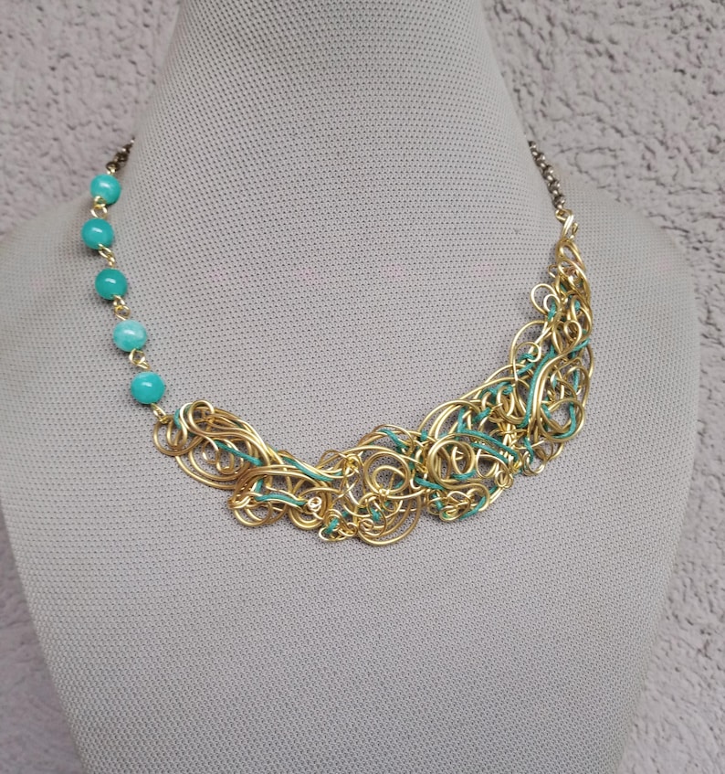 May include: A statement necklace with a gold-coloured wire design and turquoise beads. The intricate wirework is interwoven with teal accents. The necklace is displayed on a mannequin, showcasing its artistic design.