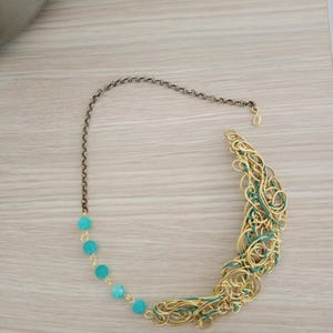May include: A gold-toned wire necklace with a turquoise bead chain and a decorative, curved pendant. The pendant features intertwined gold wire with teal accents. The necklace has a dark chain and clasp.