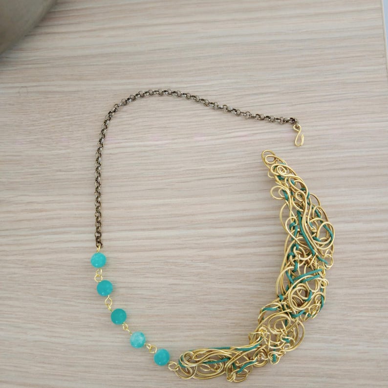 May include: A gold-toned wire necklace with a turquoise bead chain and a decorative, curved pendant. The pendant features intertwined gold wire with teal accents. The necklace has a dark chain and clasp.