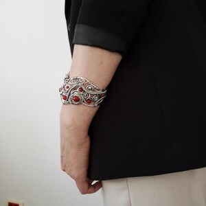 May include: A silver-toned wire cuff bracelet with red gemstone accents. The bracelet features an intricate, openwork design with circular and swirling patterns. The red stones are evenly spaced throughout the design. The bracelet is worn on a person's arm, against a black blazer.