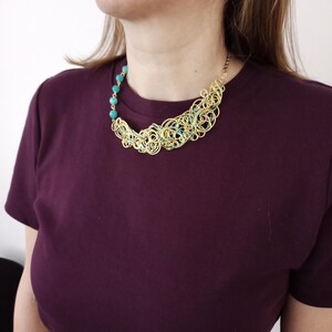 May include: A gold-coloured wire necklace featuring turquoise beads and a swirling pattern. The necklace is worn by a person in a dark purple t-shirt. The necklace has a distinctive, artistic design.