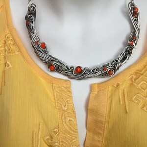 May include: A silver wire necklace with orange beads. The necklace is in the shape of a crescent moon and is on a white mannequin.