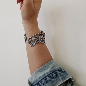 May include: A silver-toned wire cuff bracelet with red gemstone accents. The open-ended bracelet features an intricate, swirling design. The person is wearing a denim jacket with the cuff of the sleeve visible.
