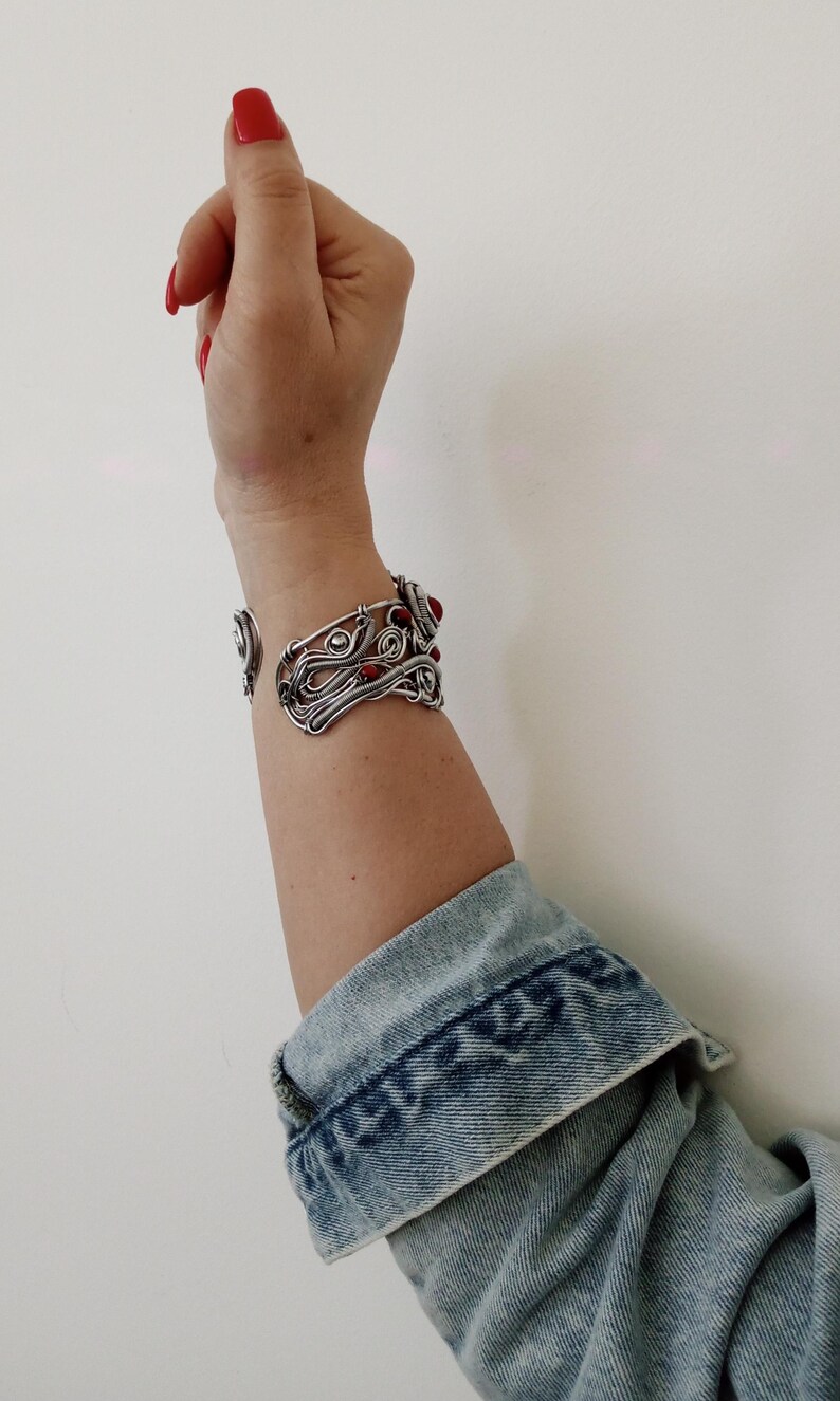 May include: A silver-toned wire cuff bracelet with red gemstone accents. The open-ended bracelet features an intricate, swirling design. The person is wearing a denim jacket with the cuff of the sleeve visible.