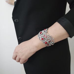 May include: A silver-toned wire cuff bracelet with red accents. The bracelet features an open design with intricate wirework and several round, red stones. The bracelet is worn on a wrist, against a black blazer.